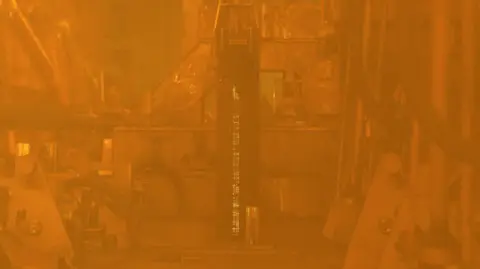 Kevin Church, BBC Plutonium is a product of reprocessing spent nuclear fuel, seen here from behind lead-lined glass. A rod of nuclear fuel is extracted, by a robotic arm, from a container. The thick, protective lead-lined glass gives a yellow tint to the scene and the nuclear fuel rod is glows because it is highly radioactive.