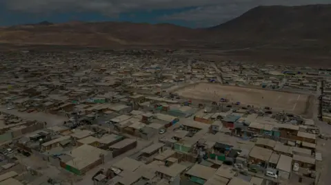 Getty Images An aerial image showing a dense collection of homes and a dirt playing field in the Flor de Poblacion encampment in Alto Hospicio, Chile. The ground looks dry and dusty and there are barren mountains in the background