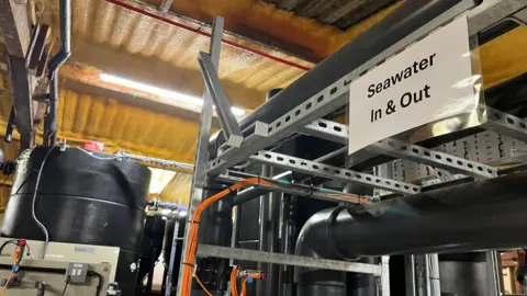 Pipes inside the facility in Weymouth. A white sign says "Seawater In and Out". There are fluorescent lights on the roof.