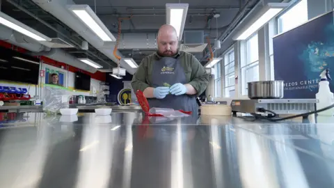 Kevin Church Well built bald man wearing a charcole grey apron, with the words "chemical kitchen", making green-coloured dumplings while wearing blue gloves.
