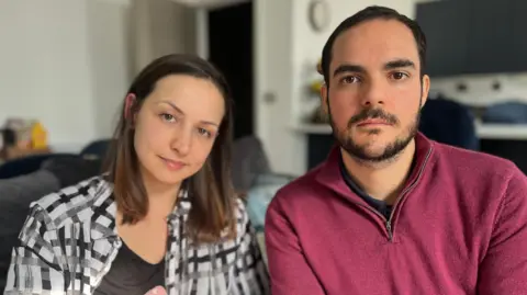 BBC A woman with brown hair and eyes wearing a checked shirt looks solemnly into the camera. She is sitting next to a man with a beard and brown eyes wearing a pink jumper with a zip.