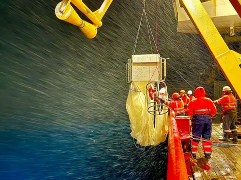 Prof Daniel J Mayor @oceanplankton Five scientists wearing high-vis orange jackets and dark trousers on board the Sir David Attenborough polar ship. They are working with a fishing net equipped with 9 closing nets and has a 1 × 1 m mouth.  There is a large yellow crane above them.